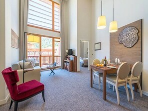 A spacious living area with a red chair, a dining table with chairs, a TV, and a large window with blinds.