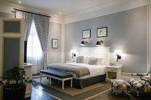 Quarto com cama grande, mesas de cabeceira, um banquinho, uma cadeira e um vaso de planta.