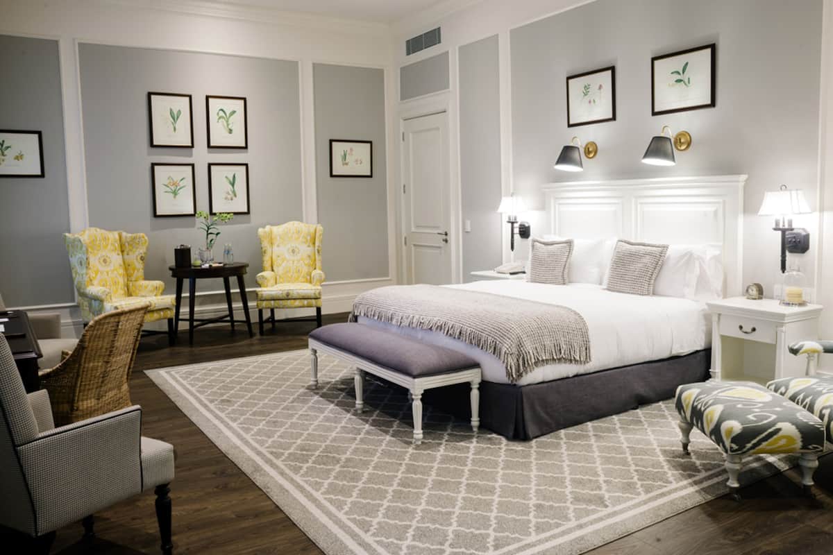 A bedroom with a large bed, two bedside tables, a nightstand, a bench, two chairs, and a small table with a vase.