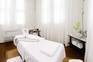 Um quarto limpo e claro com uma cama, uma mesa de massagem, uma mesinha com toalhas, uma pia e uma planta.