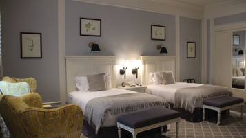 A hotel room with two beds, a large window, a chair, and a bedside table with a lamp.