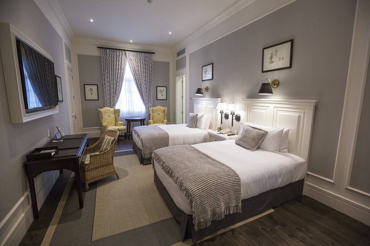 A hotel room with two beds, a desk, a chair, a TV, and a window with curtains.