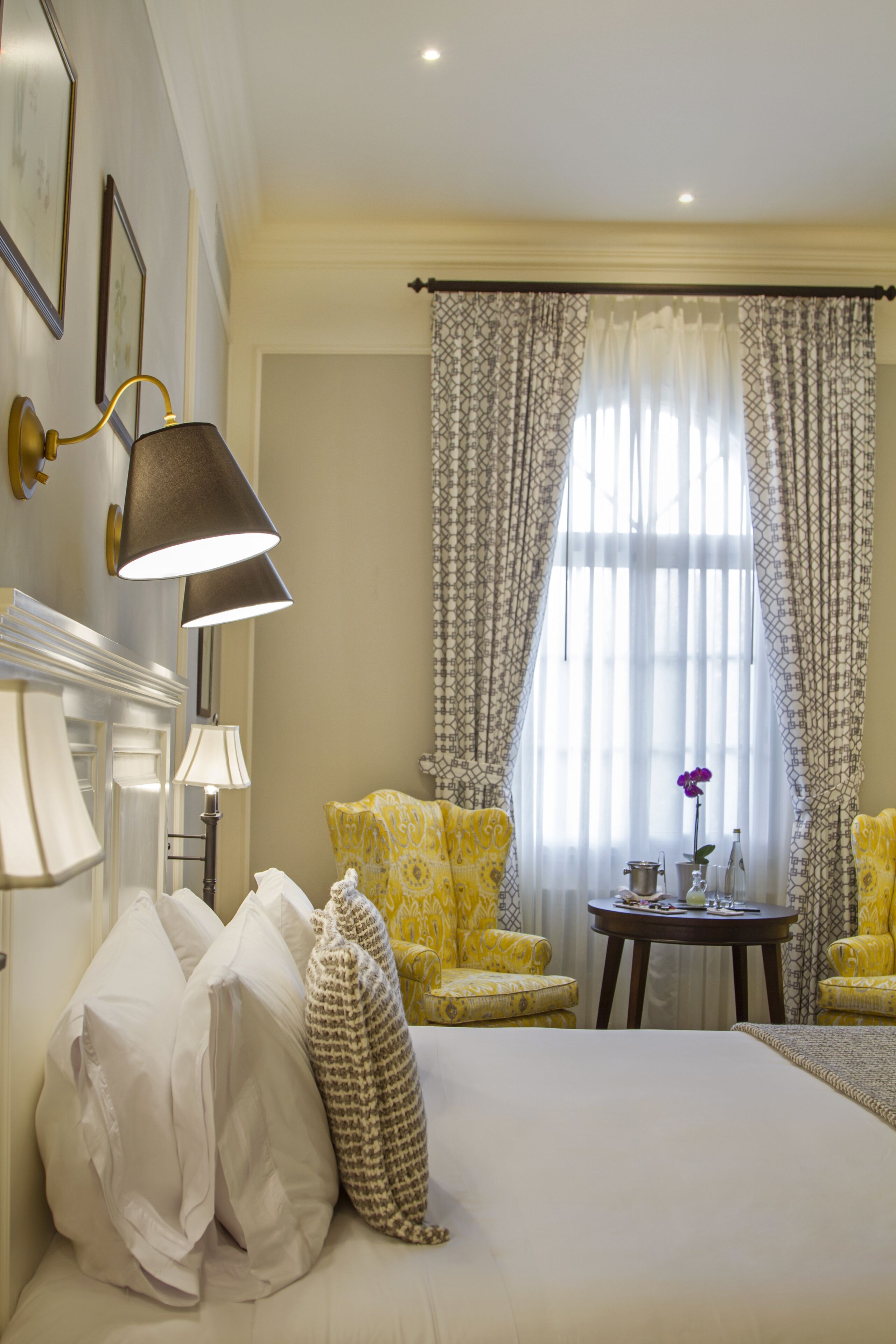A hotel room with a bed, two yellow armchairs, a small table with a vase, and a lamp.