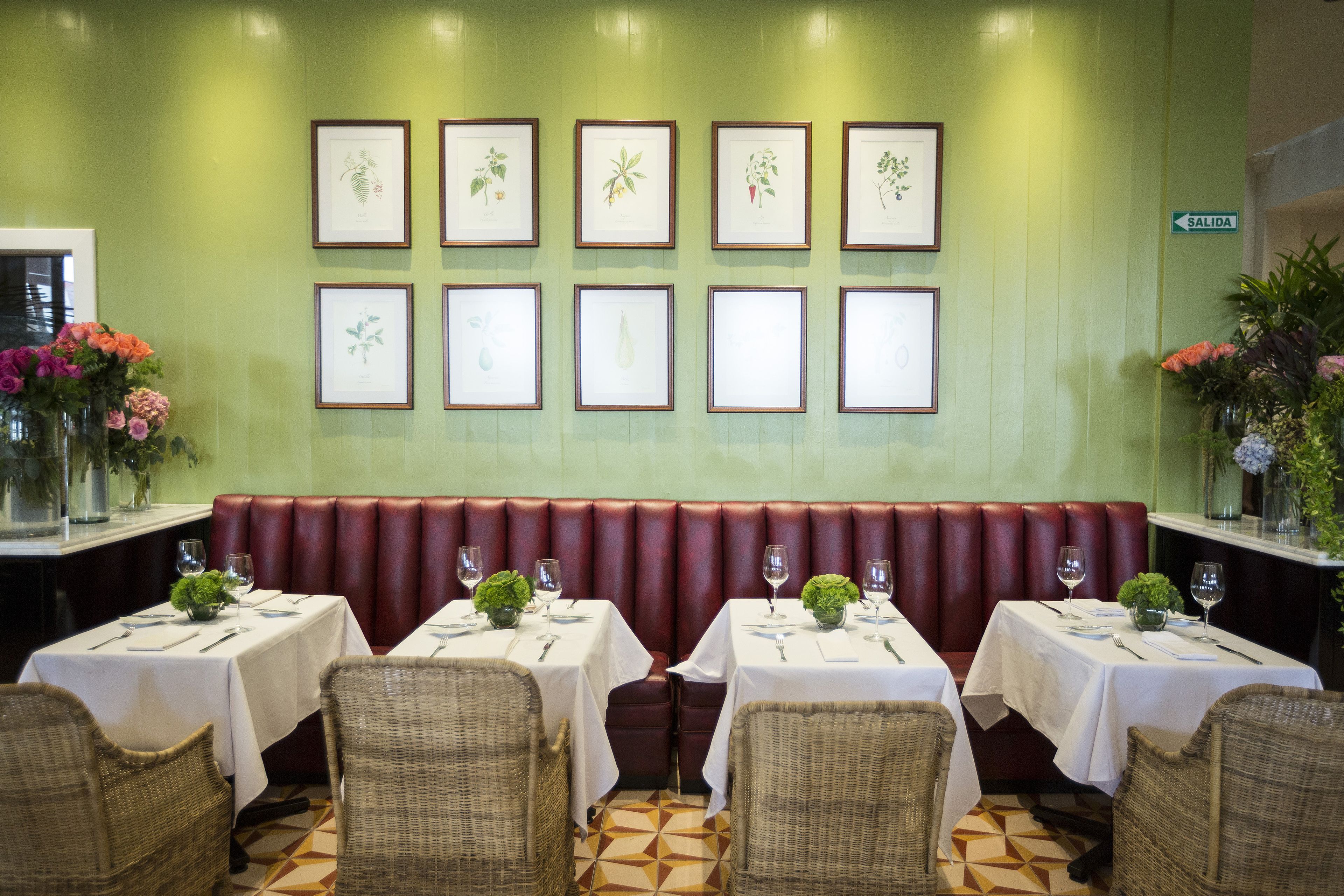 Un restaurante con una pared verde, impresiones botánicas enmarcadas, una zona de asientos con bancos rojos y mesas dispuestas con manteles blancos y cristalería.