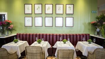 Un restaurante con una pared verde, impresiones botánicas enmarcadas, una zona de asientos con bancos rojos y mesas dispuestas con manteles blancos y cristalería.