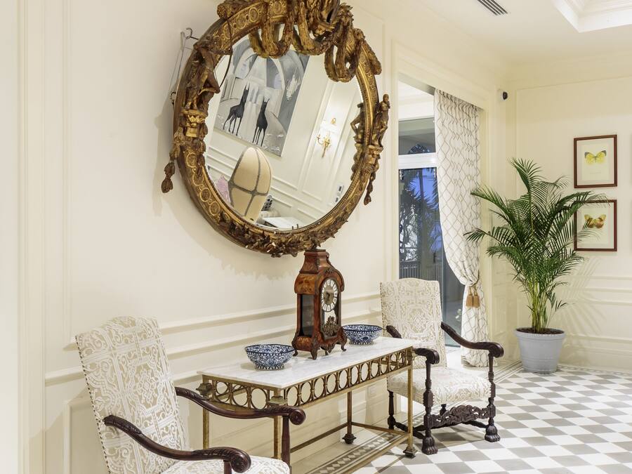 Un suelo de tablones encalados, un espejo ornamentado y dos sillones tapizados en una habitación con pared blanca y una planta en maceta.