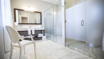 Un baño moderno con cabina de ducha de vidrio, suelo de mármol blanco, un espejo grande y una silla blanca.