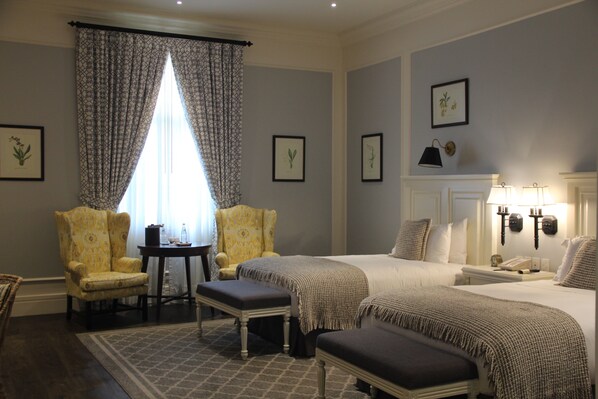 Una habitación de hotel con dos camas, una zona de estar con una silla y una mesa, e impresiones botánicas enmarcadas en las paredes.