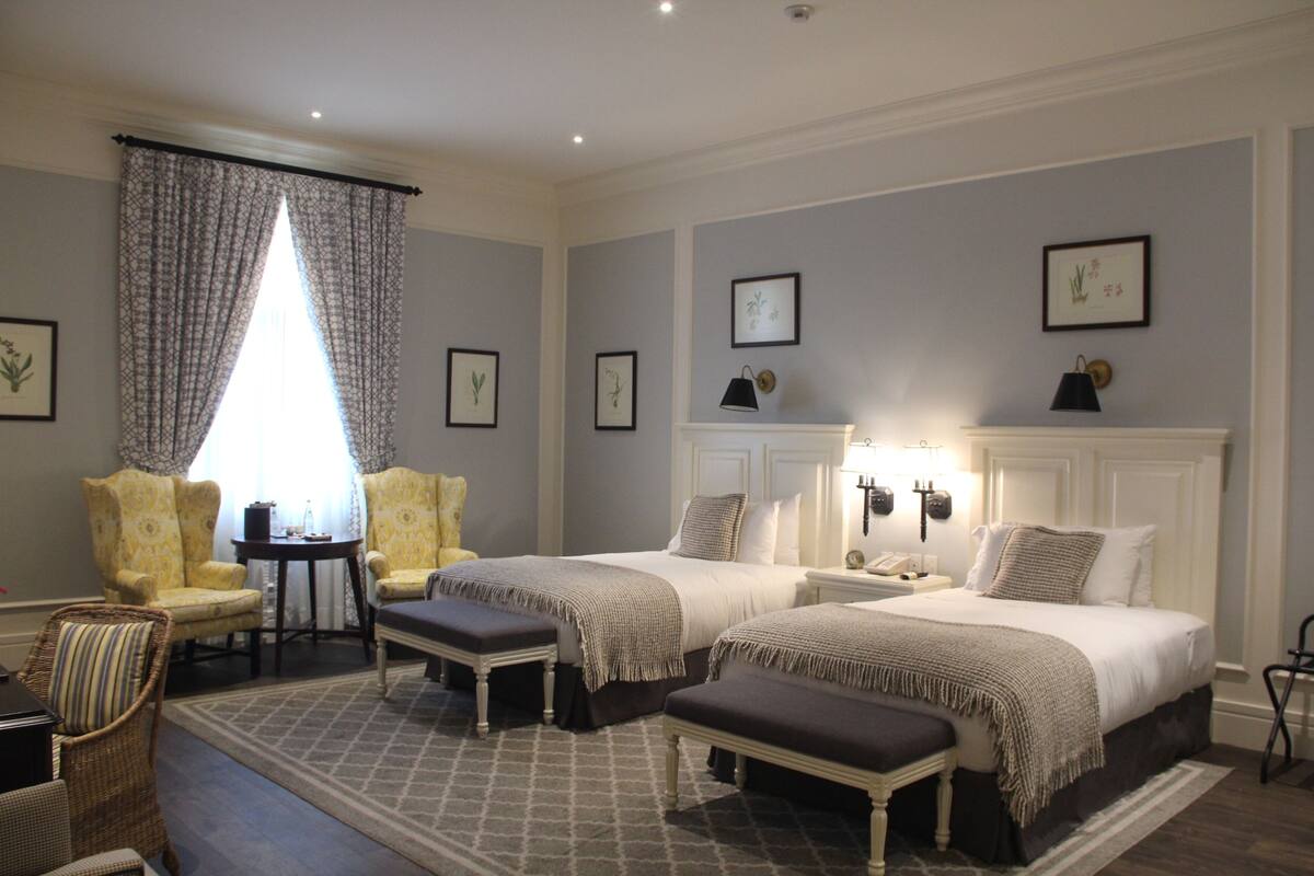 A hotel room with two beds, a sitting area with chairs, a small table, and a large window with curtains.