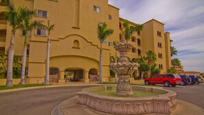 Un edificio con una fuente en el patio, palmeras y vehículos estacionados.