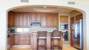 A kitchen with wooden cabinets, a stainless steel refrigerator, a microwave, and two wicker bar stools. - Las Mananitas LM 3 Villa 3501 3 Bedroom Villa By Seaside Los Cabos (San José del Cabo)
