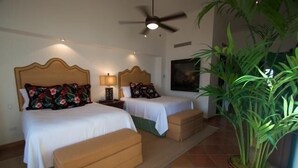 Habitación de hotel con dos camas, ventilador de techo, mesita de noche, lámpara y una planta grande en maceta.