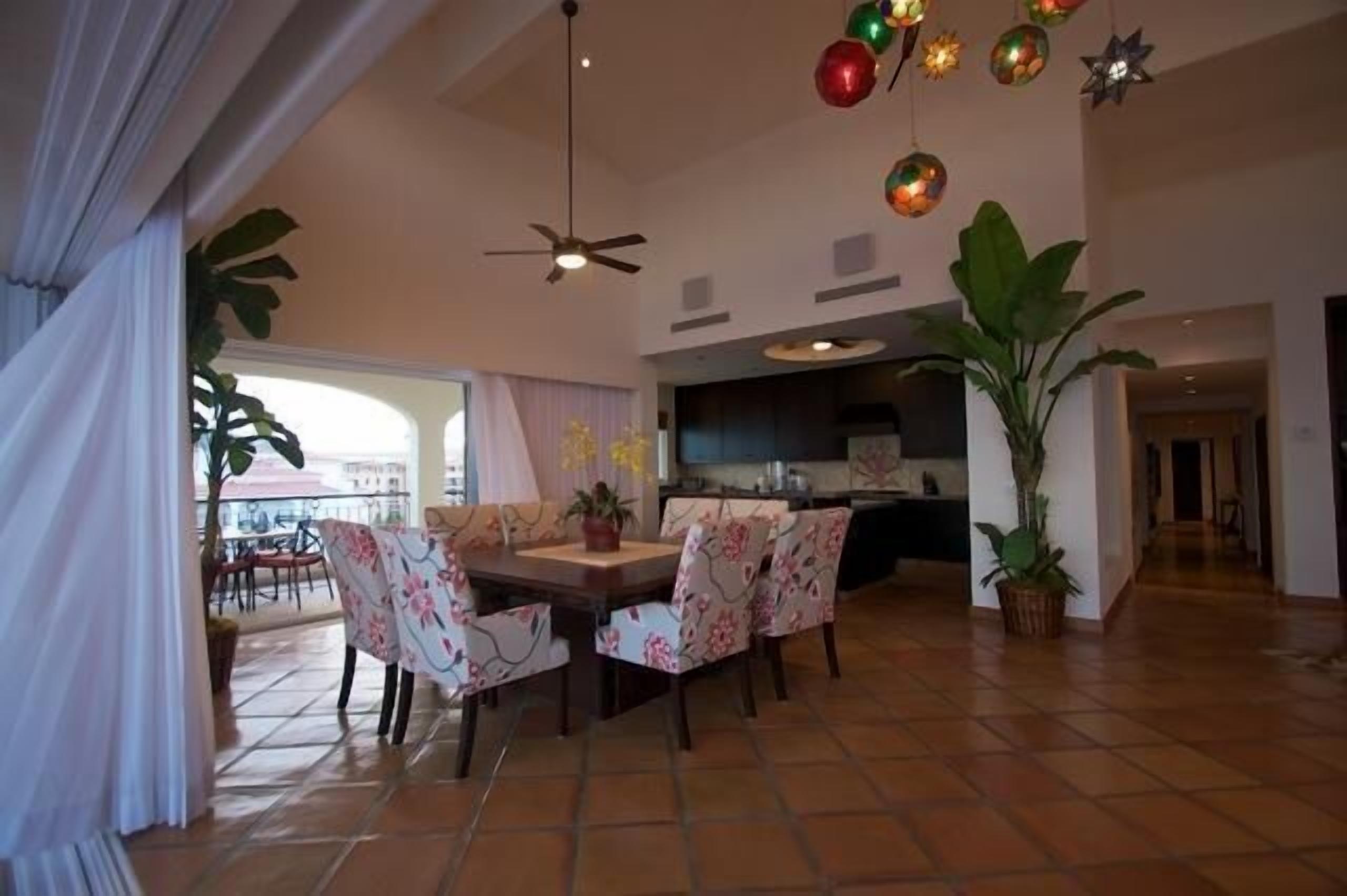 Une salle à manger spacieuse avec un grand ventilateur de plafond, une cuisine à l’arrière-plan et de grandes plantes en pot.