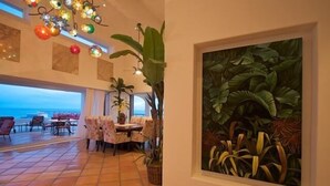 Un comedor con vistas al océano, un cuadro de una planta tropical grande y un techo con luces colgantes.
