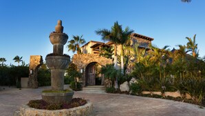 A courtyard with a stone fountain, palm trees, and a building with a stone facade and arched entrance.