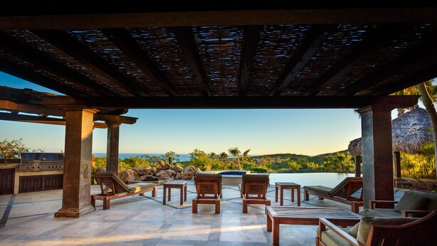 A covered outdoor seating area with lounge chairs and a grill, overlooking a pool and scenic landscape.