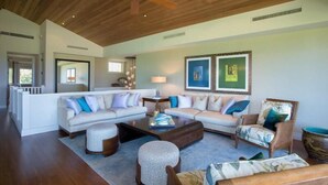 A spacious living room with a large sectional sofa, a coffee table, and a patterned ottoman. There are two side tables with lamps, and the room has a wooden ceiling. - Palm Villa 126B (Kailua-Kona)