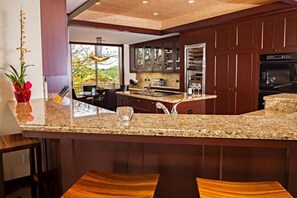 A modern kitchen with granite countertops, dark wooden cabinets, and a large island with bar stools. - 115D Waiulu Street Hualalai (Kailua-Kona)