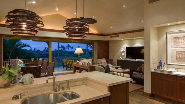A modern kitchen with a large island, a dining area with a TV, and a view of palm trees. - 129B Hualalai (Kailua-Kona)
