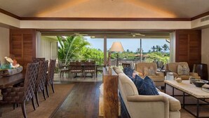 A spacious living area with a sofa set, dining table, and chairs. A large window offers a view of lush greenery and palm trees. - 129B Hualalai (Kailua-Kona)