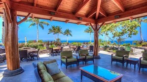 A covered outdoor seating area with a pool table, green sofas, and chairs, overlooking a beach with palm trees and the ocean.