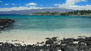 A beach with clear turquoise water, a rocky foreground, and a distant view of a resort with palm trees and a mountain in the background. - Fairways at Mauna Lani 1305 (Kamuela)