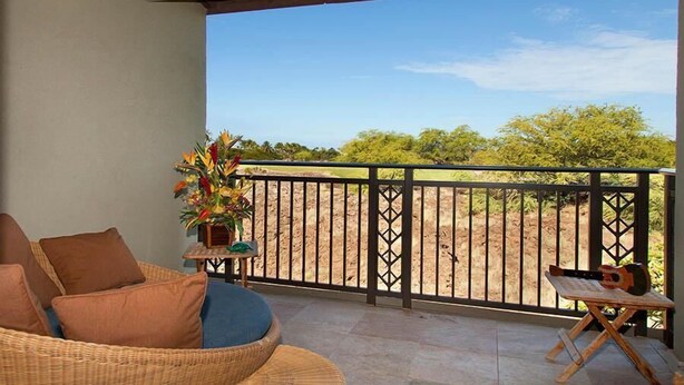 A balcony with wicker furniture, a small table with a guitar, and a view of trees and sky. - Fairways at Mauna Lani 1104 (Kamuela)