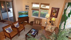 A living room with a TV, two sofas, a coffee table, and a large window with a view of greenery. - Fairways at Mauna Lani 1104 (Kamuela)