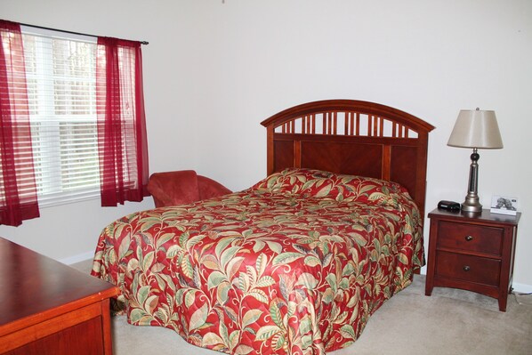 A bedroom with a wooden bed, a nightstand with a lamp, a desk, and a chair.