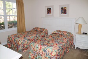 A bedroom with two beds, a window with curtains, a bedside table with a lamp, and two framed pictures on the wall.