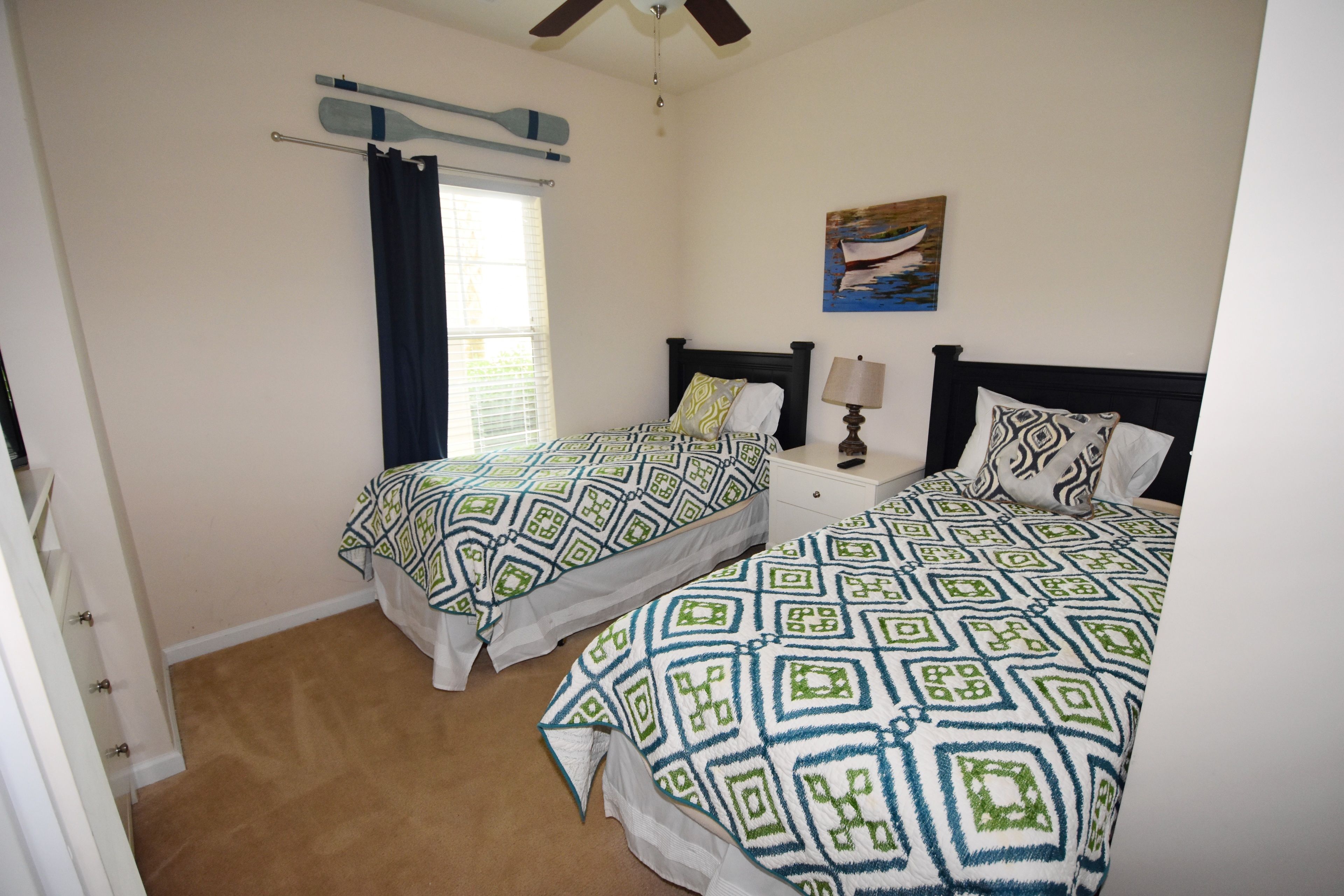 A bedroom with two beds, a ceiling fan, a window with curtains, a bedside table with a lamp, and a painting on the wall.