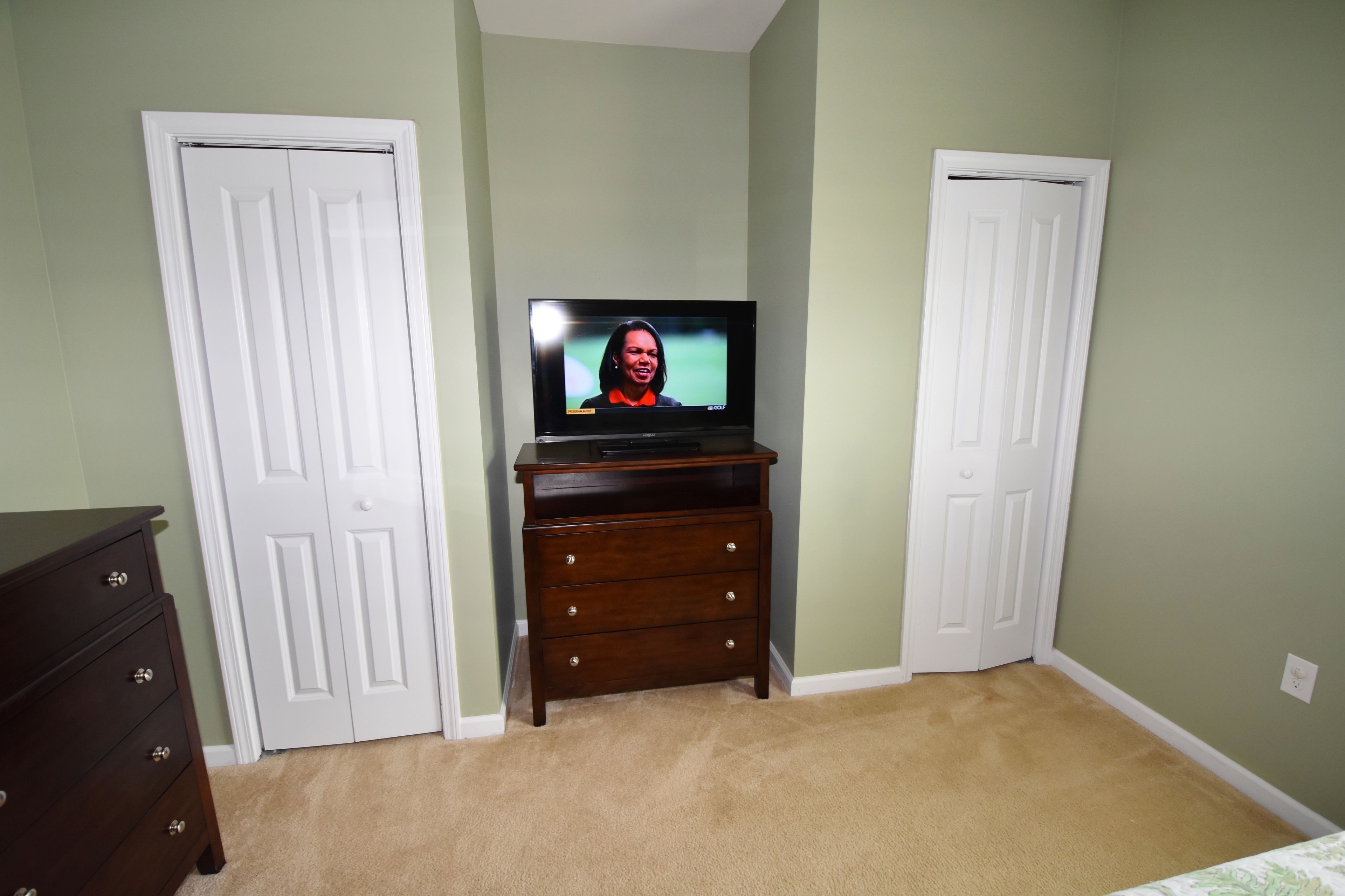 Een kamer met een televisie op een standaard, twee witte deuren, een donker kastje en een beige vloerkleed.