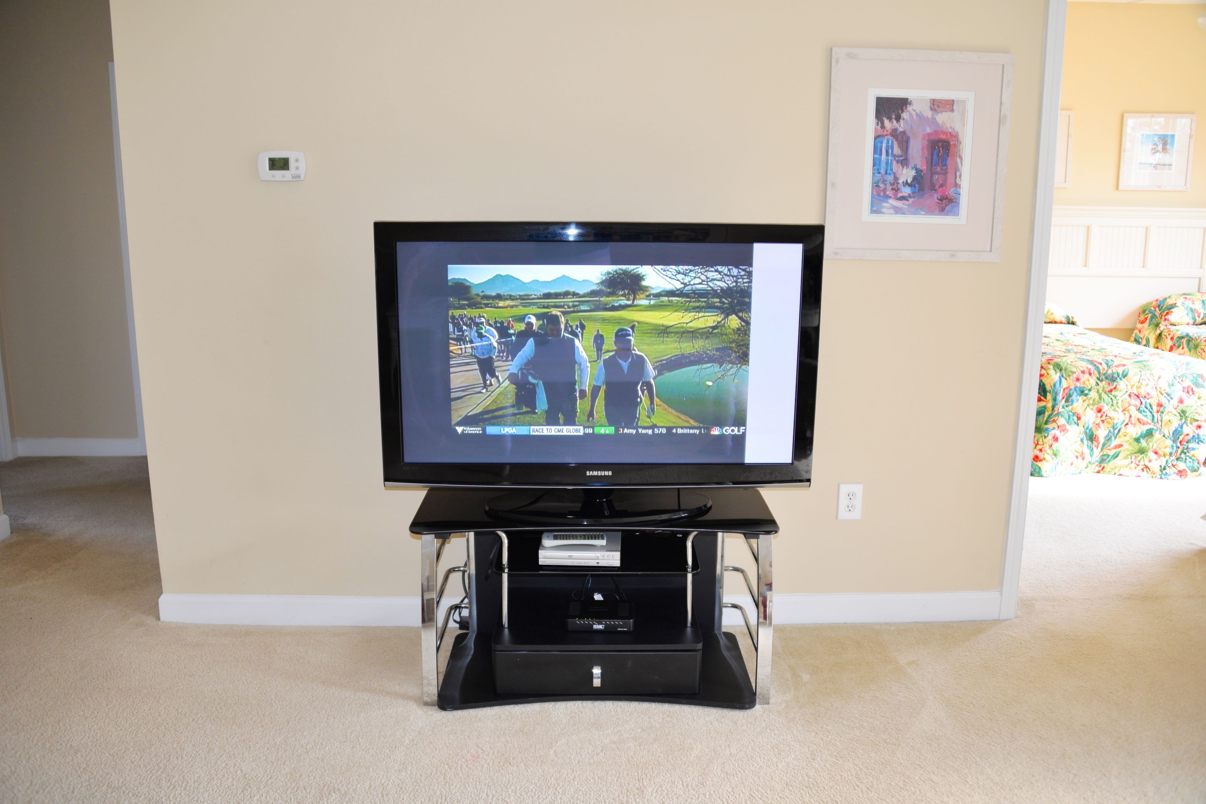 A television on a black stand in a room with beige walls, carpet, and a bed with floral bedding.