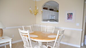 A dining area with a round wooden table surrounded by six white chairs, a chandelier, and a kitchen visible through an archway. - 1523 The Havens (North Myrtle Beach)