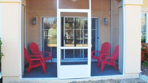 Un área de estar techada al aire libre con dos sillas rojas y una mesa, delimitada por un ventanal con marco blanco.
