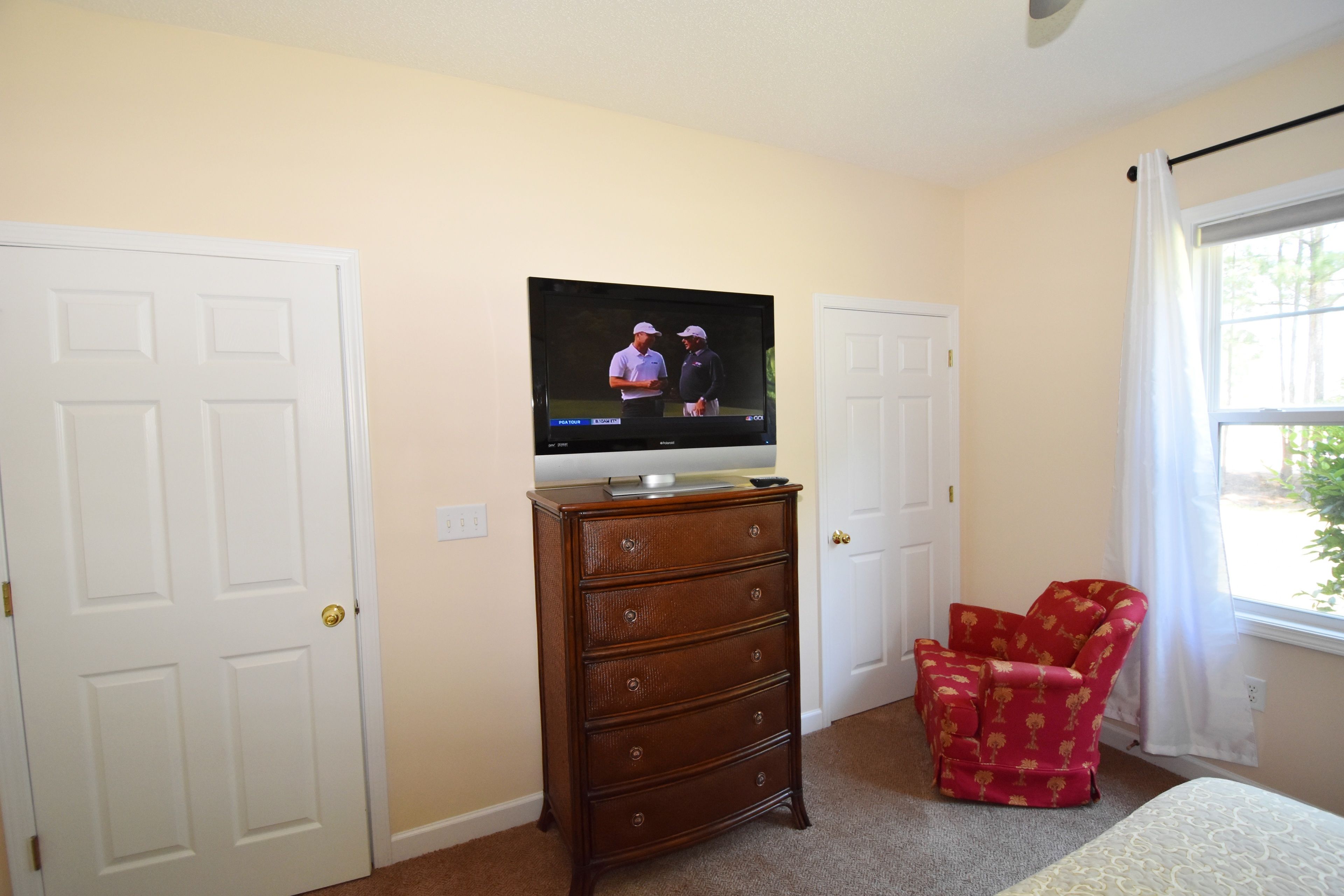 Un dormitorio con una televisión sobre un aparador, una silla roja y una ventana con cortinas.