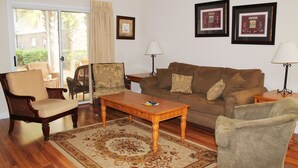 A living room with a sofa, two armchairs, a wooden coffee table, and a patterned rug. There are two lamps, a door leading outside, and three framed pictures on the wall. - 614 Willow Bend (North Myrtle Beach)