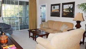 A living room with a beige sofa, a coffee table, and a lamp. There are two framed pictures on the wall and a window with curtains. - 734 The Havens (North Myrtle Beach)