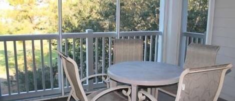 A covered patio with a round table and four chairs, a white railing, and a view of trees through large windows.