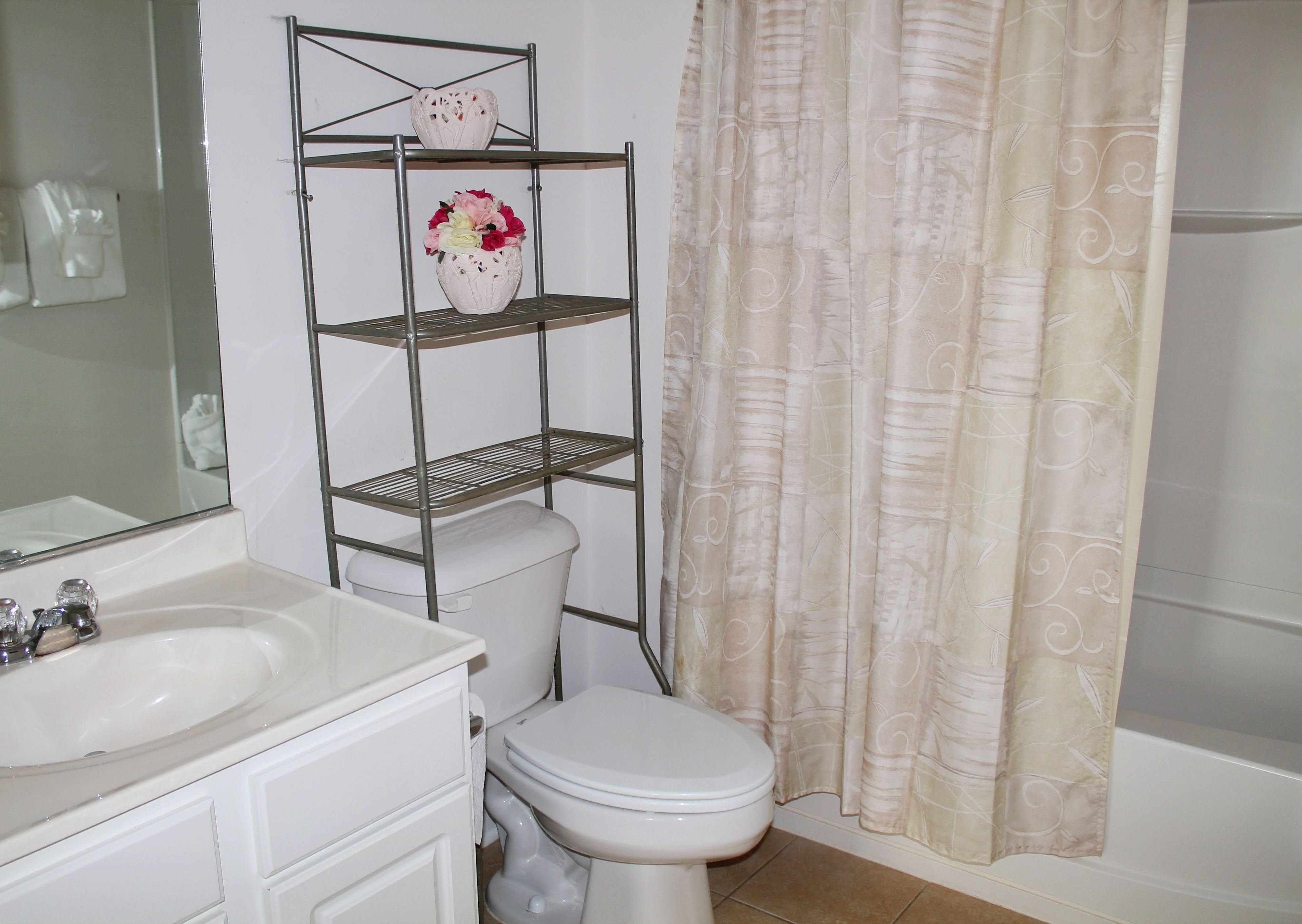 A bathroom with a white sink, toilet, and shower curtain. There is a metal shelf unit with a vase of flowers on the top shelf.