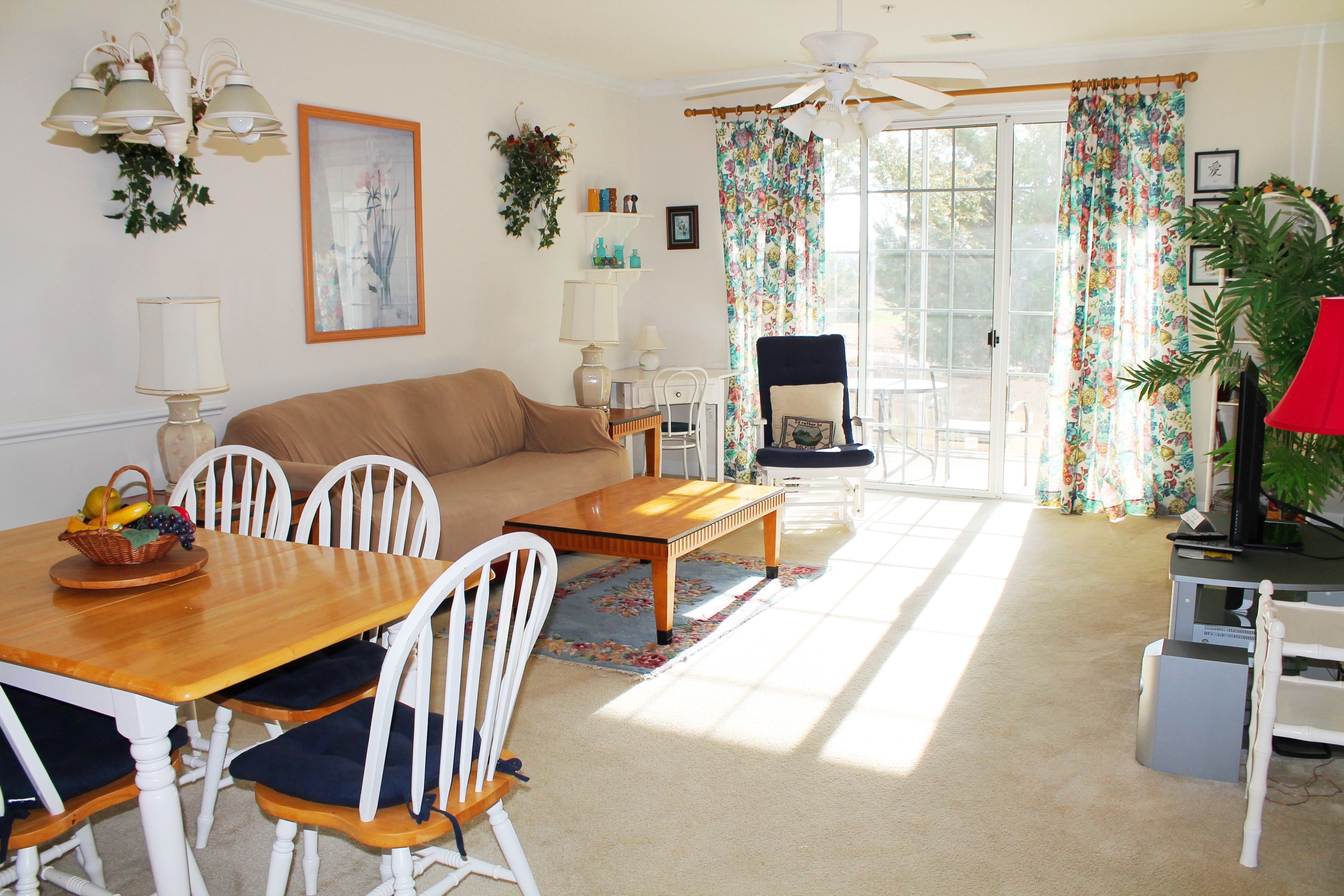A living room with a wooden dining table, white chairs, a sofa, and a ceiling fan.