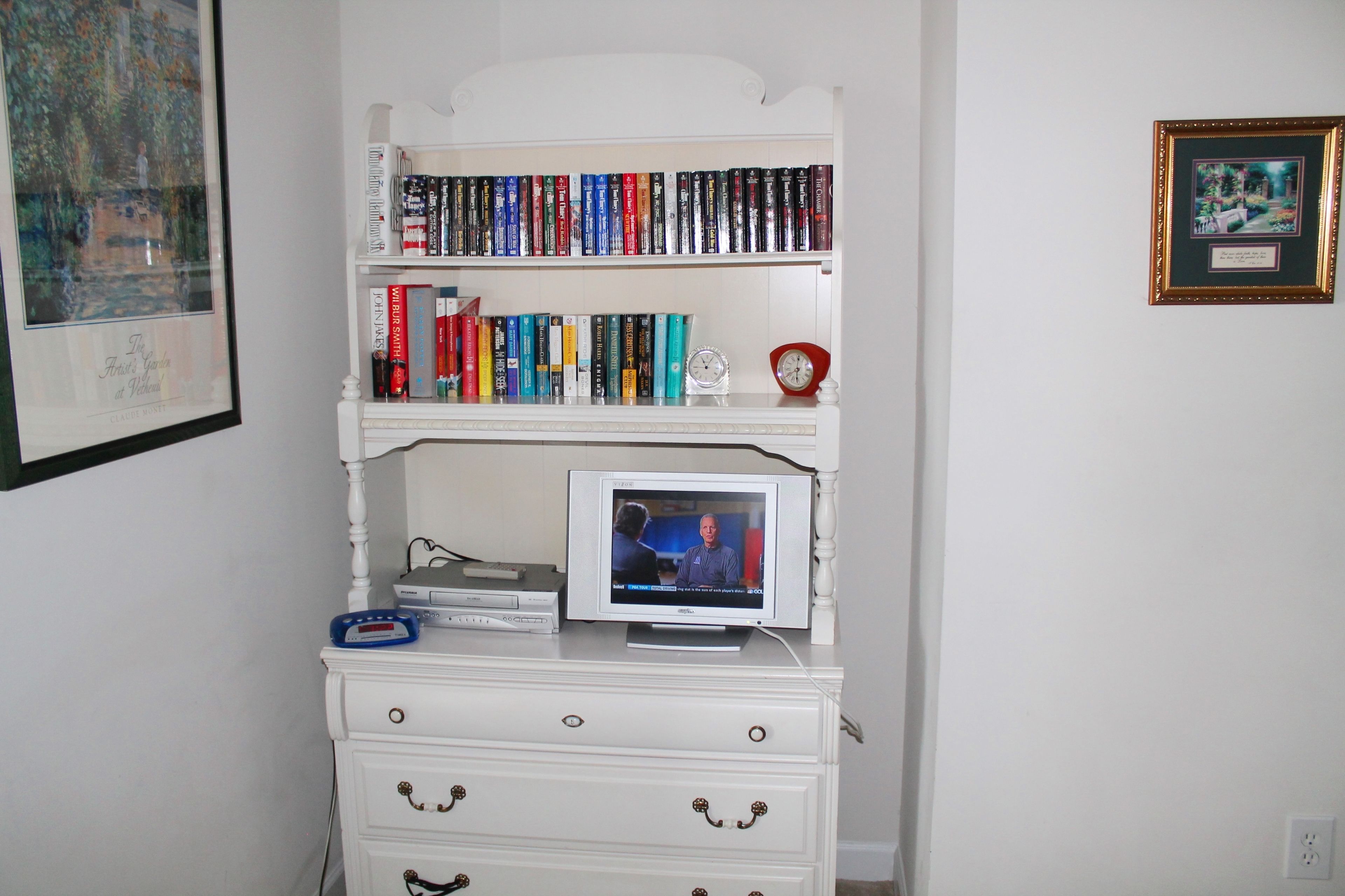 A white dresser with a television on top, a bookshelf with books, and a framed picture on the wall.