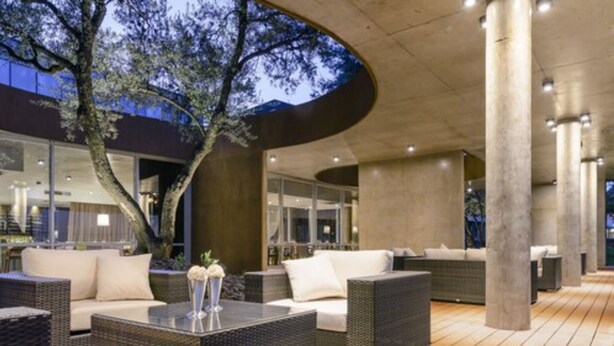 An outdoor lounge area with wicker furniture, a glass enclosure, and a tree. - Epic Hotel Villa de Merlo (Merlo)