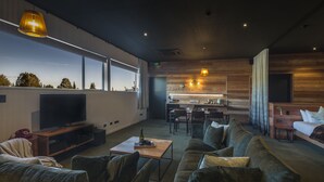 A modern living room with a large TV, comfortable seating, a kitchen area, and a view of the outdoors. - The Cinema Suites (Te Anau)
