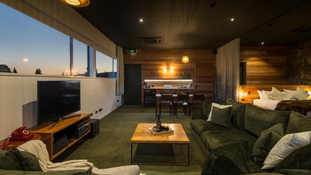 A modern living room with a TV, sofa, and coffee table. A kitchen is visible in the background with a dining area. - The Cinema Suites (Te Anau)