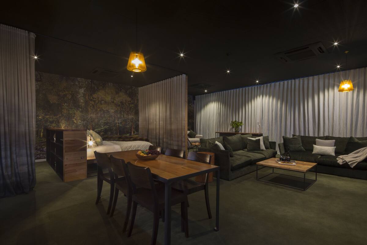 A modern living room with a wooden dining table, a sofa, and a coffee table. There is a bed with a headboard and a curtain. The room has a textured wall and a ceiling with recessed lighting.