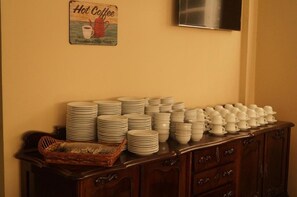 Montaje de bufé con platos y recipientes apilados, una cesta de mimbre y un letrero en la pared que dice 'Café Caliente'.