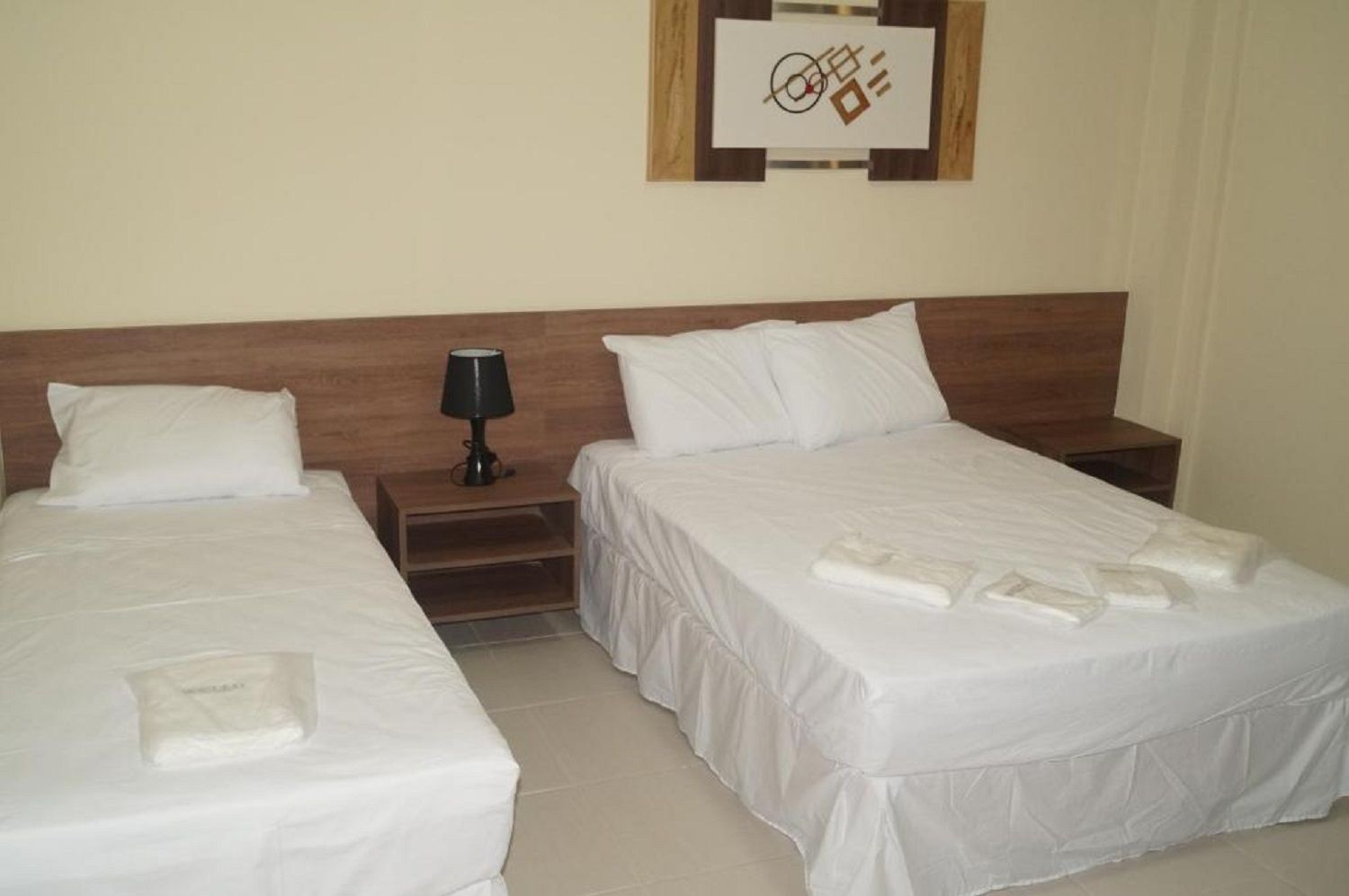 Quarto de hotel com duas camas, uma mesa de cabeceira com abajur e uma obra de arte na parede.