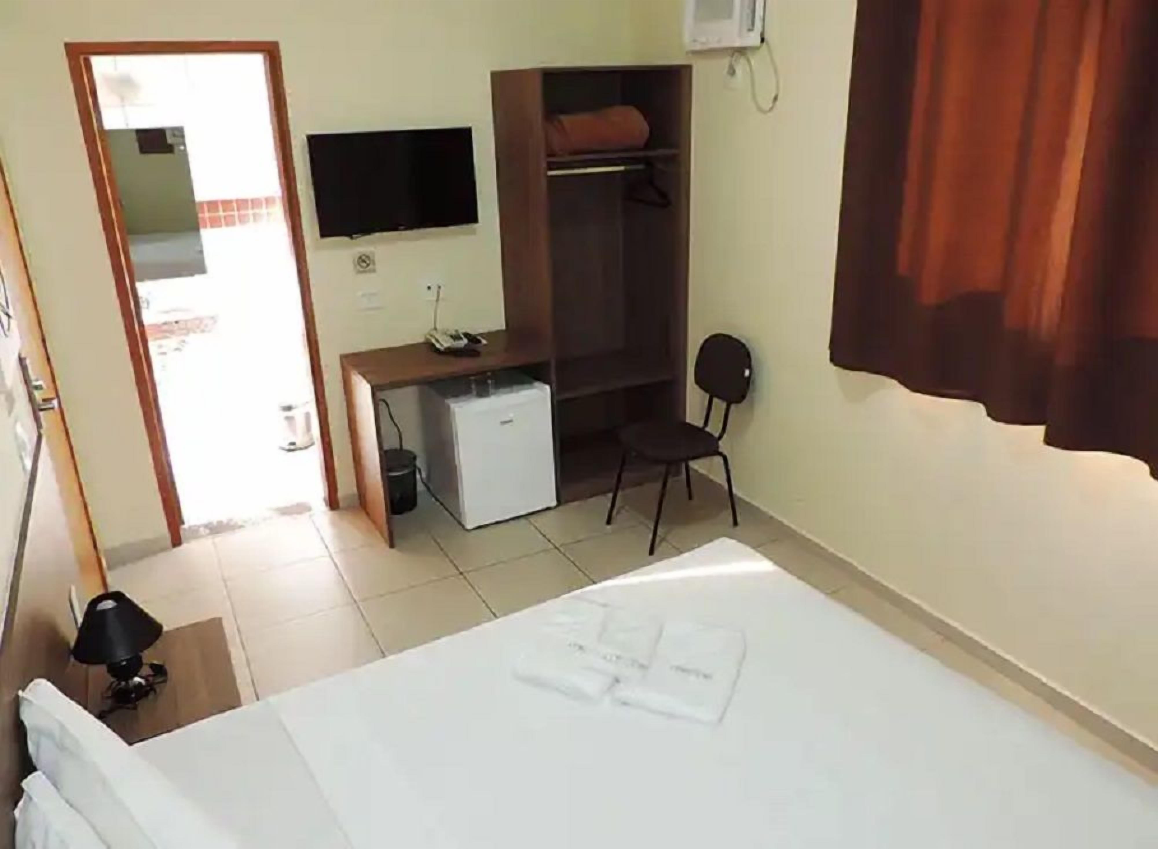 A hotel room with a bed, a desk with a computer, a small fridge, a chair, and a TV mounted on the wall.