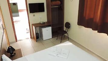 A hotel room with a bed, a desk with a computer, a small fridge, a chair, and a TV mounted on the wall.
